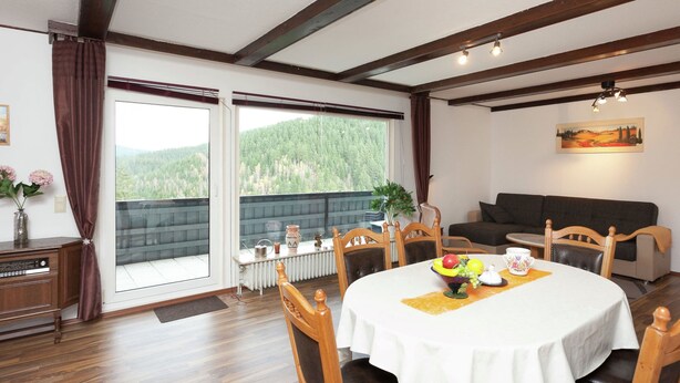 Ein Essbereich mit rundem Tisch, Holzsesseln, einer Couch und Blick auf einen Wald durch große Fenster.