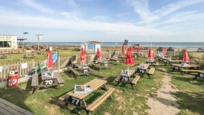 Un área de picnic junto a la playa con mesas numeradas y sombrillas rojas, un pequeño edificio identificado como 'NIS BAY', y vista al mar de fondo.