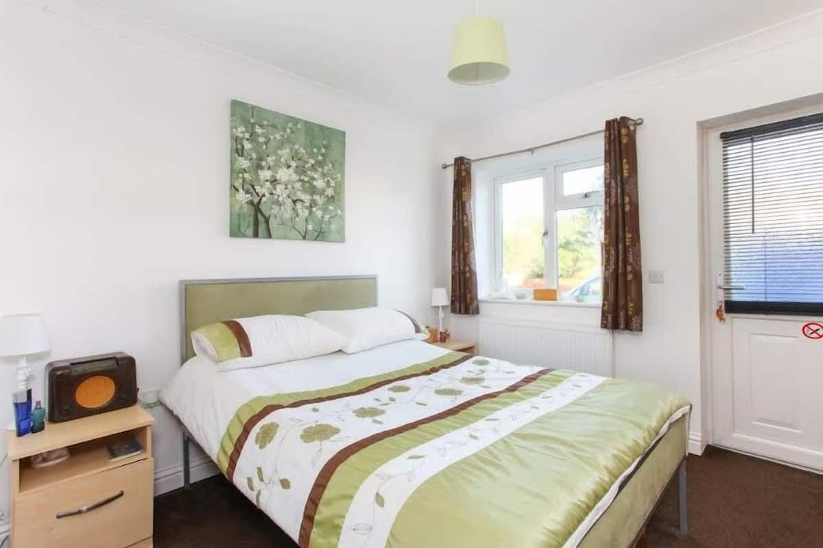 Un dormitorio con cama, mesita de noche, radio, un cuadro, una ventana con cortinas y una puerta con letrero.