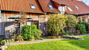 Een tweelaags houten huis met een schuin rood dak, een tuin met diverse planten en een geplaveid pad.