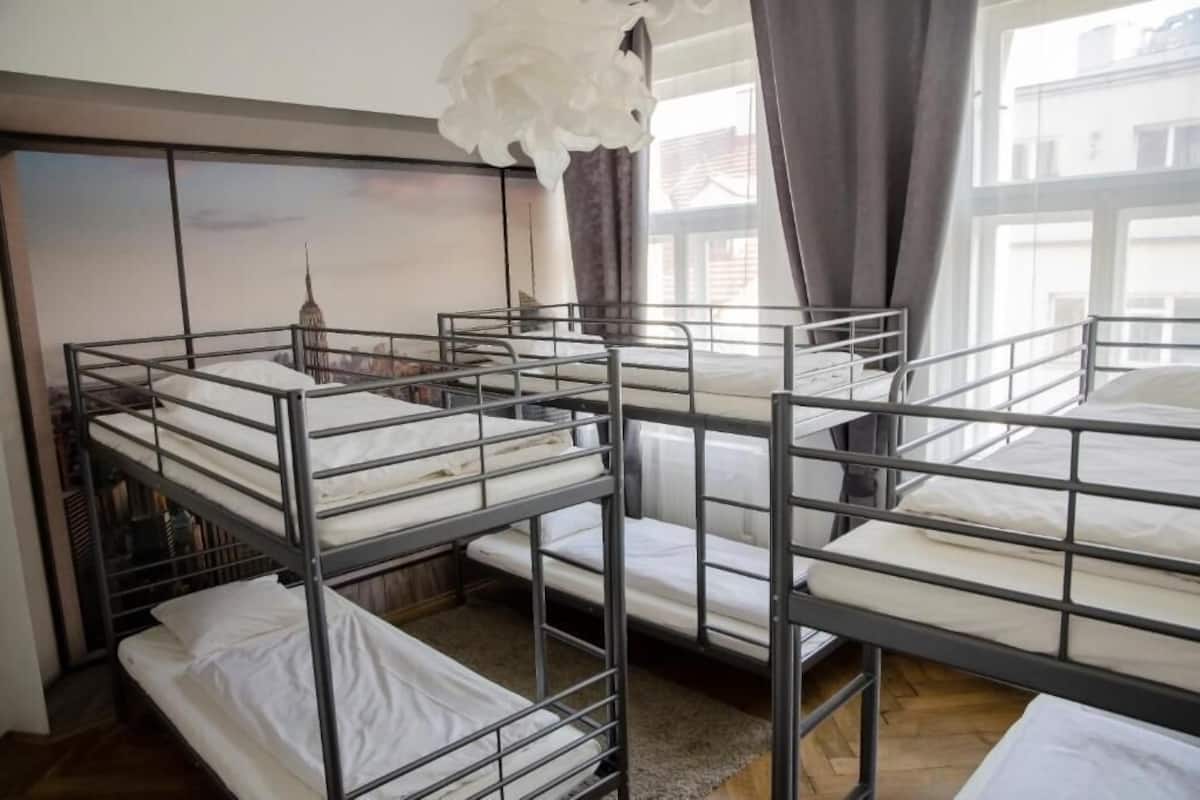 A dormitory room with bunk beds, a large window with curtains, and a view of a cityscape.
