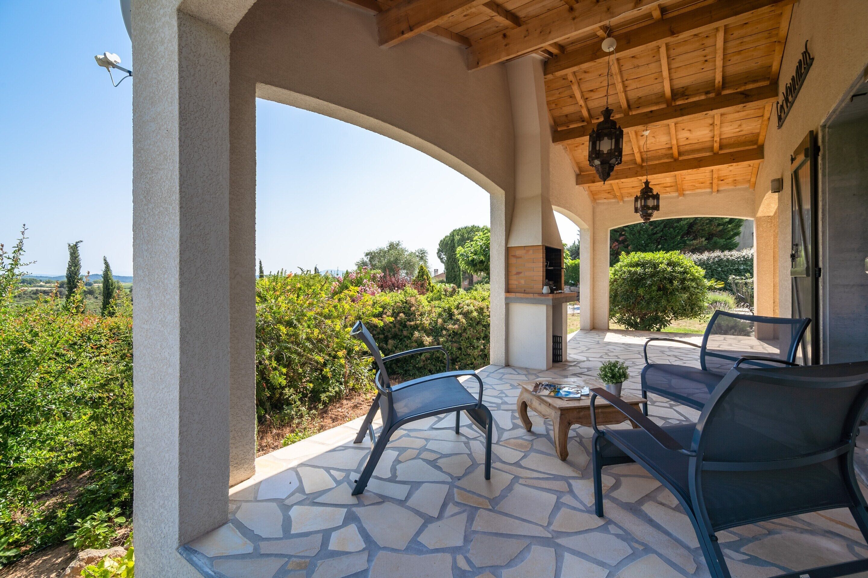 A covered patio with stone flooring, two chairs, a small table, and a view of greenery and a building in the background.