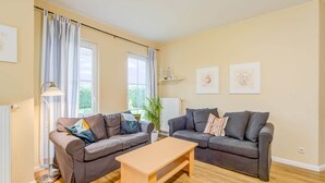Un salon avec deux canapés gris, une table basse en bois et un lampadaire. Il y a deux fenêtres avec des rideaux, un radiateur et une plante en pot.