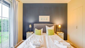 Une chambre à coucher avec un grand lit, deux tables de chevet, une fenêtre avec des rideaux et un mur foncé orné d’un tableau encadré.