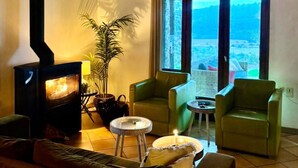 A cozy living room with a wood-burning stove, two green armchairs, a small table, and a view of the outdoors through large windows. - Charming French Villa With Pool (Cebazan)