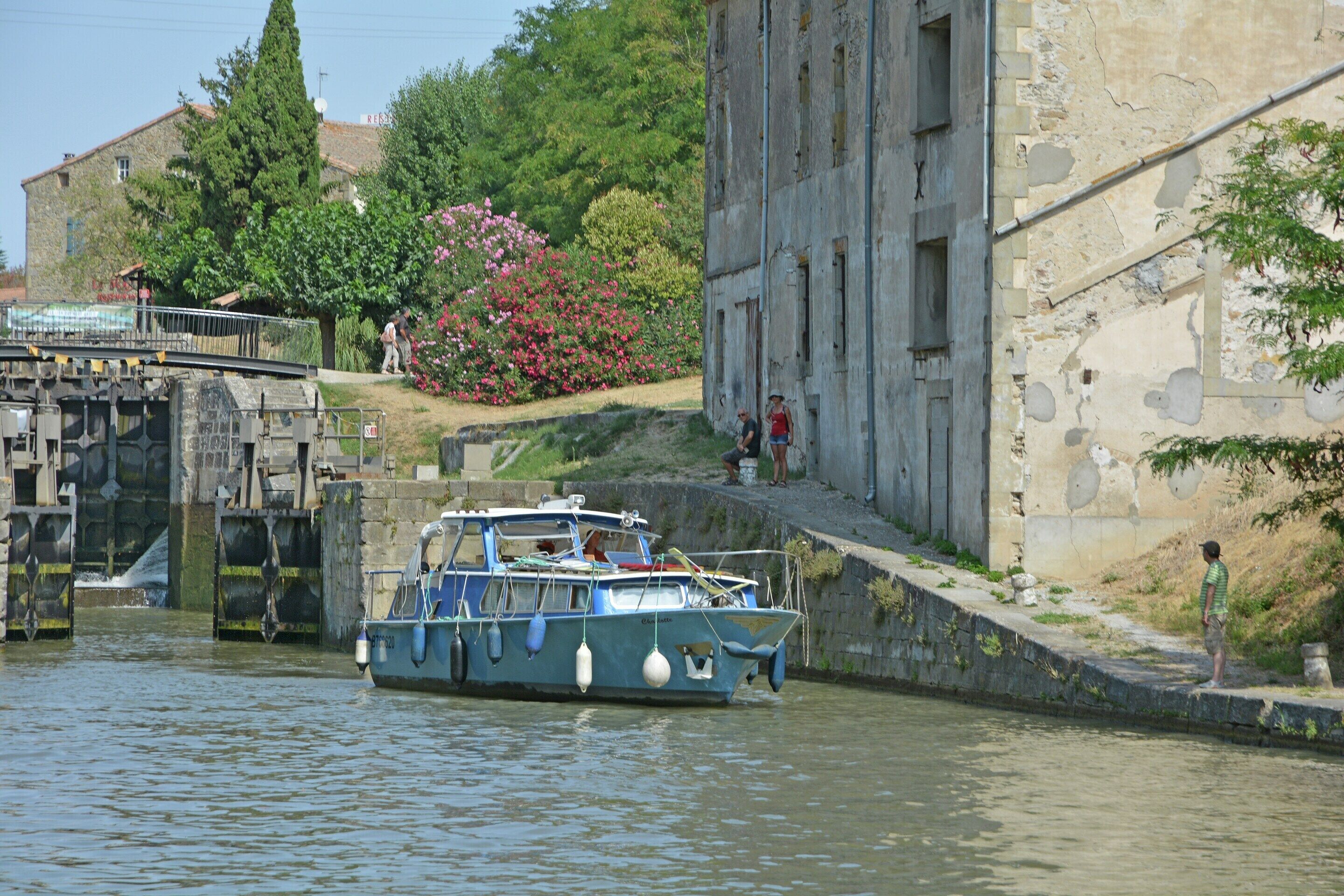 A boat docked at a canal with a stone building and greenery in the background.