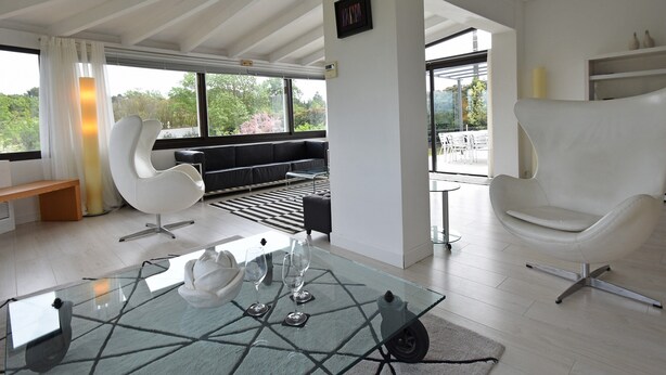 A modern interior with a glass coffee table, white chairs, a sofa, and large windows offering a view of greenery. - Villa in Malves Near Carcassonne Pool (Malves-en-Minervois)