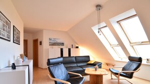 Ein modernes Wohnzimmer mit einer schwarzen Ledersofa, zwei schwarzen Ledersesseln, einem Holztisch und einer weißen Kommode. Zwei Dachfenster sorgen für natürliches Licht.