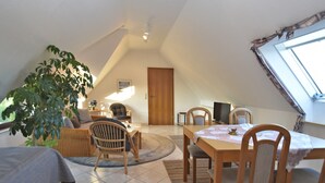 A cozy, well-lit attic bedroom with a bed, a desk, a chair, a television, and a large window.
