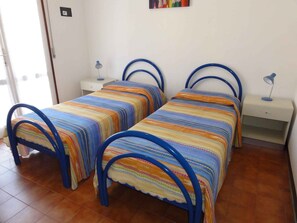 Een kamer met twee bedden, elk met een blauw metalen frame en kleurrijk gestreept beddengoed. Er zijn nachtkastjes met lampen en een klein plankje.