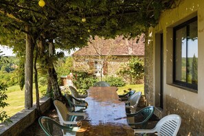 An outdoor dining area with a long wooden table and plastic chairs, surrounded by greenery and a stone building in the background. - Holiday Home in Dordogne With Pool (Thenon)