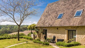 Une maison en pierre avec un toit en pente, deux fenêtres, une terrasse avec une table et des chaises, et un arbre au premier plan.