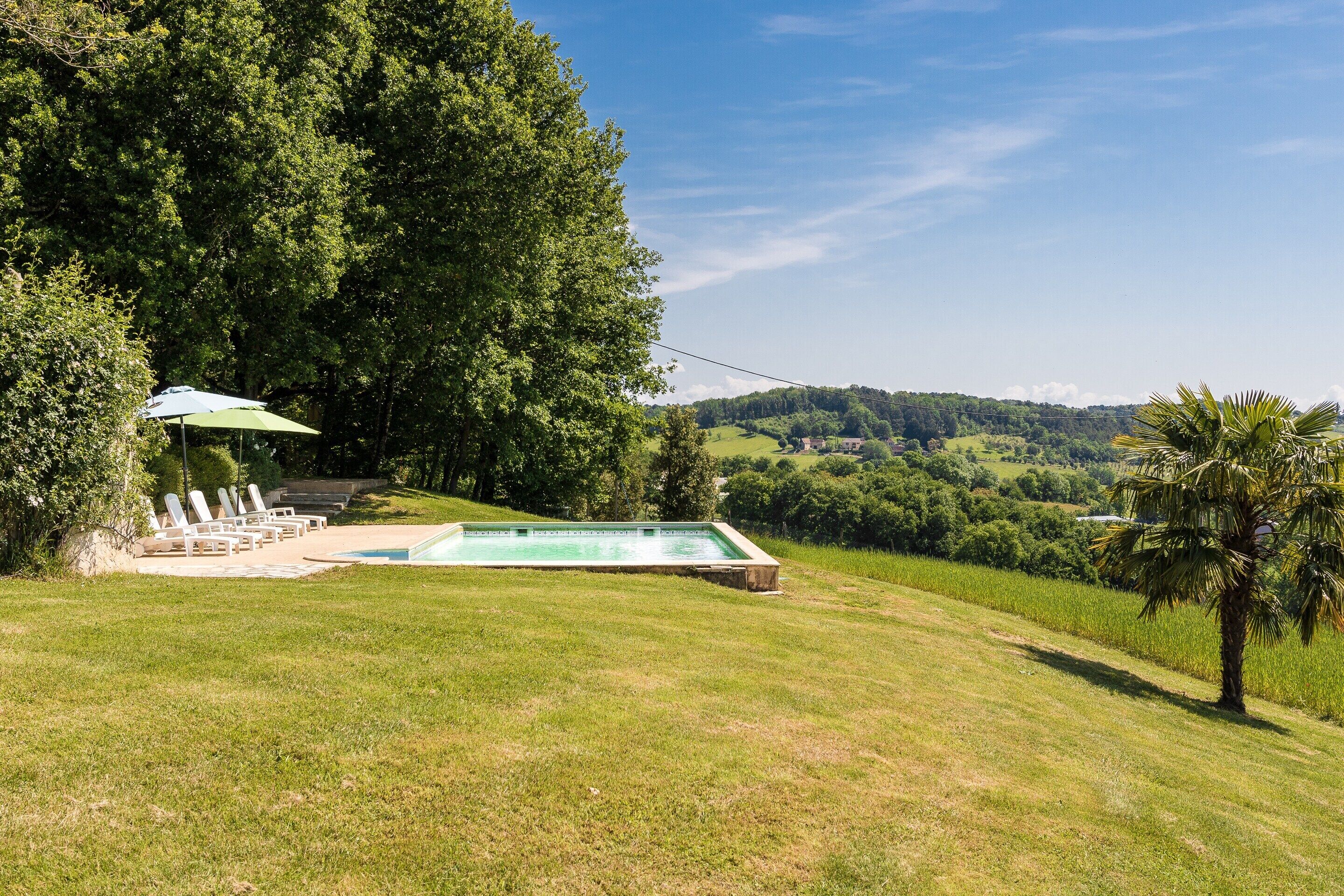 A swimming pool with lounge chairs and an umbrella, surrounded by lush greenery and hills in the background.