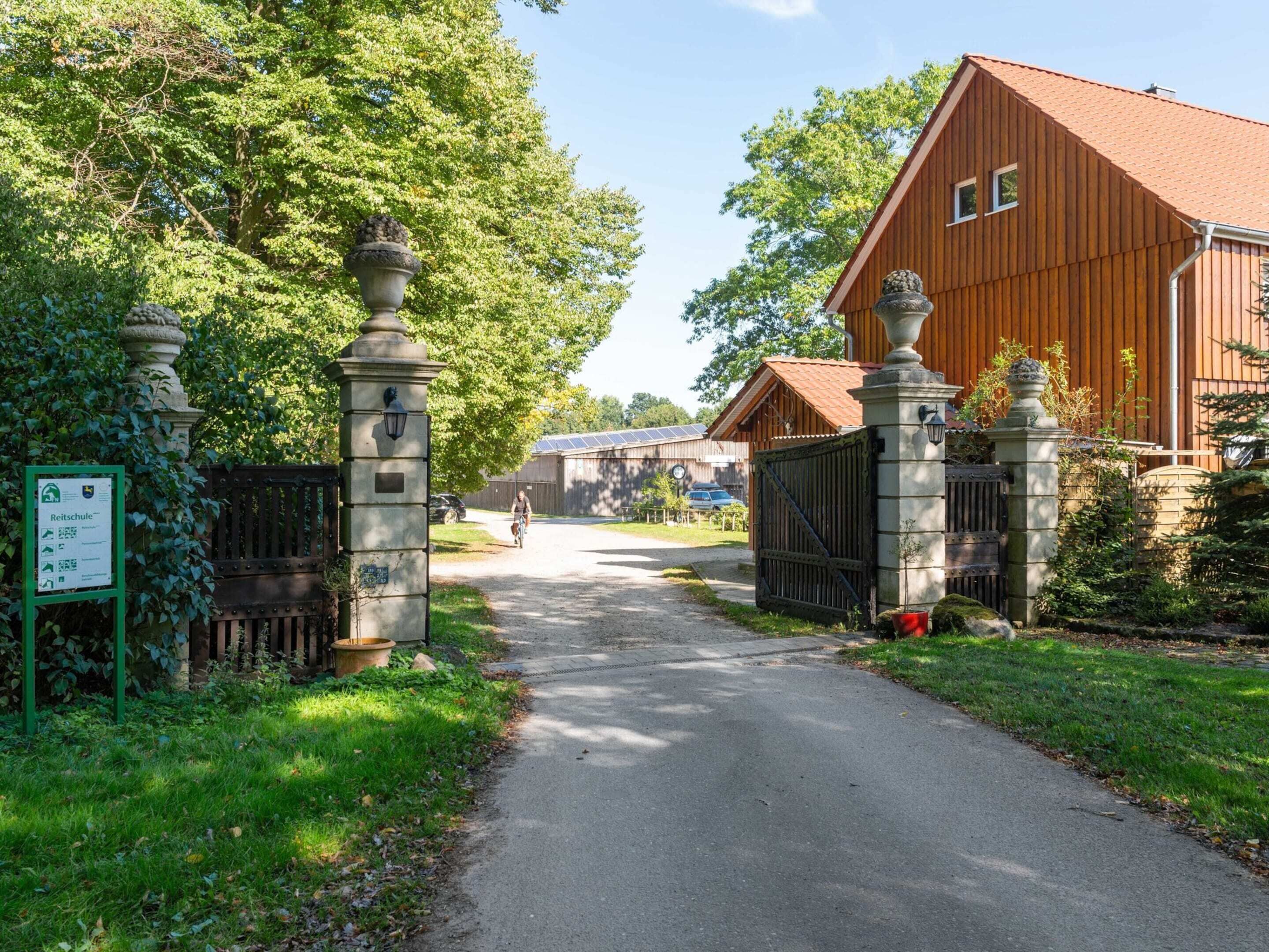 A rural road leading to a wooden building with a red roof, surrounded by greenery and stone pillars.