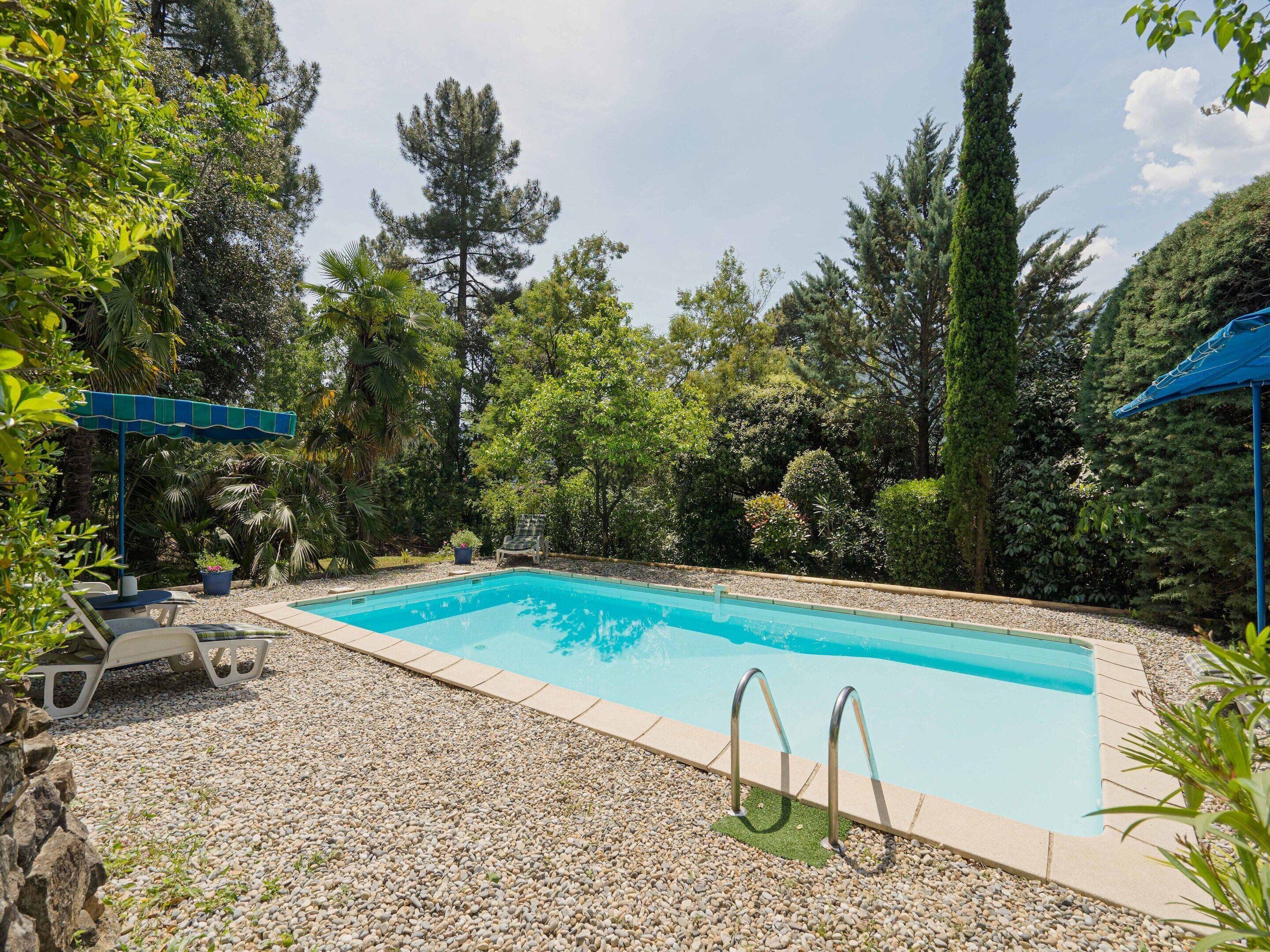 A rectangular swimming pool with a pebble border, surrounded by lush greenery and trees.