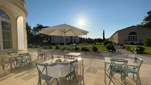 Un patio al aire libre con mesas y sillas, un gran paraguas y un jardín cuidado con una fuente.