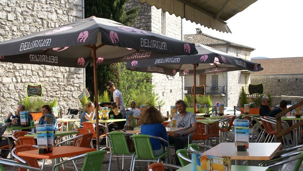 Ein Straßencafé mit Tischen, Stühlen und Sonnenschirmen, im Hintergrund eine Steinmauer.