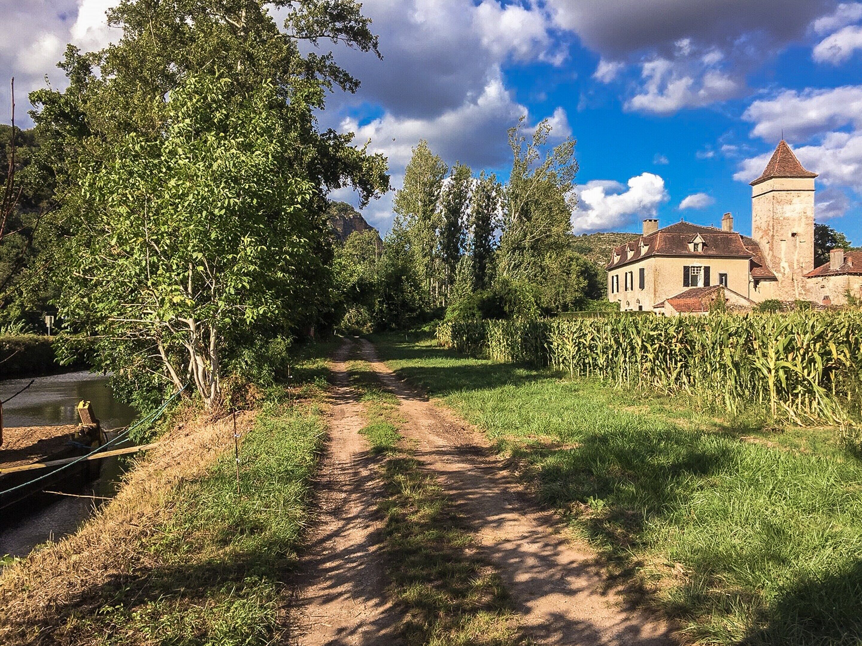 Un sentier rural menant à un bâtiment historique doté d’une tour, entouré d’arbres et d’un champ de maïs.