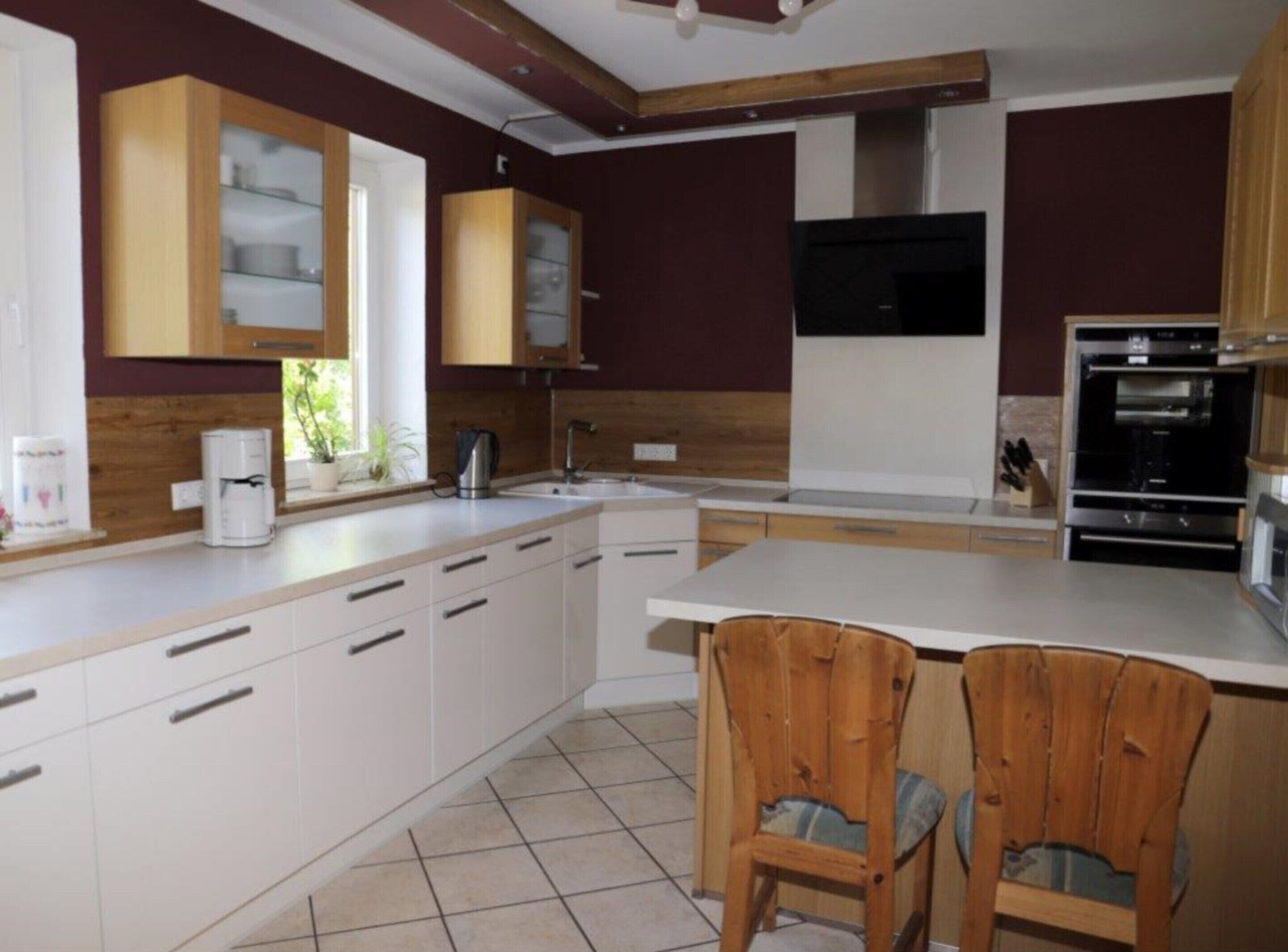 A modern kitchen with white cabinets, a large island, wooden chairs, and built-in appliances.