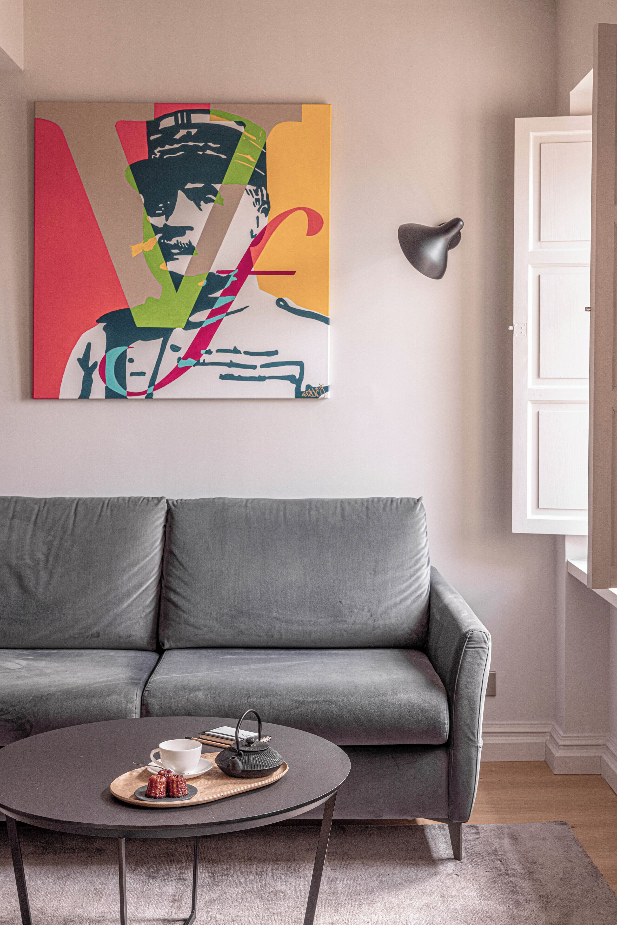 A modern living room with a grey sofa, a round coffee table with a tray, a teapot, and a cup. A colorful abstract painting is on the wall.
