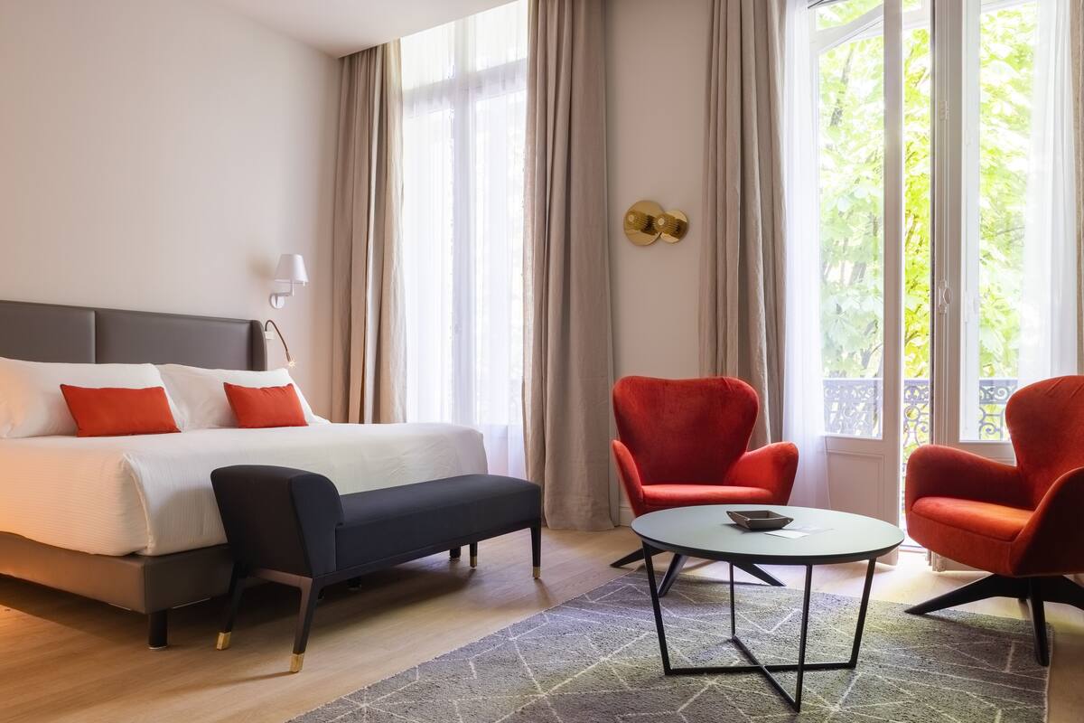 Quarto de hotel com cama, um banco preto, uma mesa redonda e duas poltronas vermelhas.