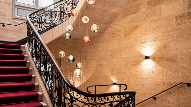 A grand staircase with red carpeted steps, ornate black metal railings, and hanging decorative lights. - Villas Foch (Bordeaux)