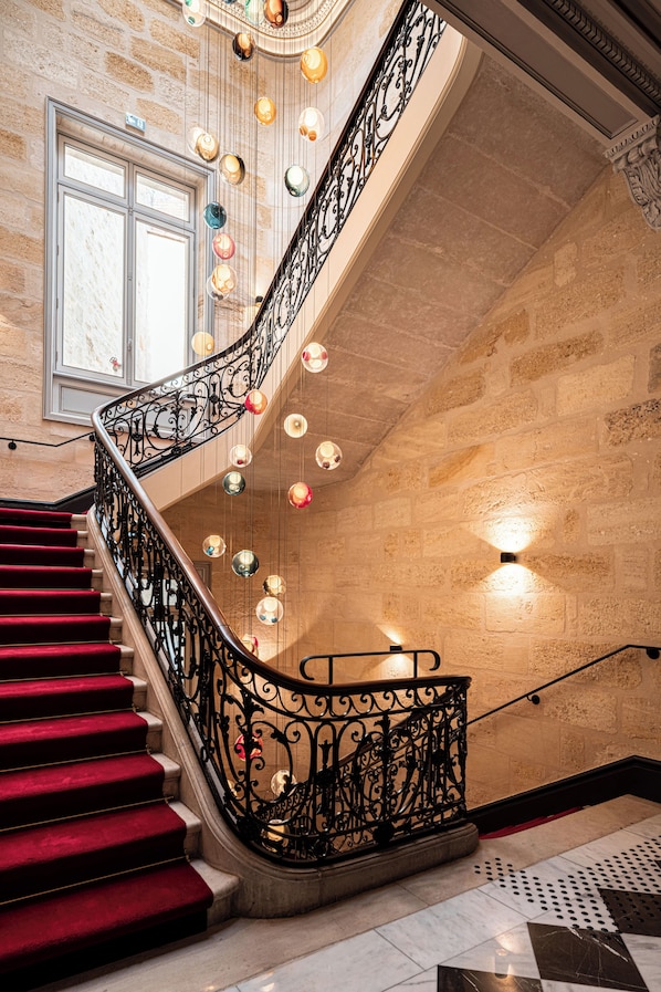 Una imponente escalera con alfombra roja, barandales de metal negro ornamentados y luces decorativas colgantes.