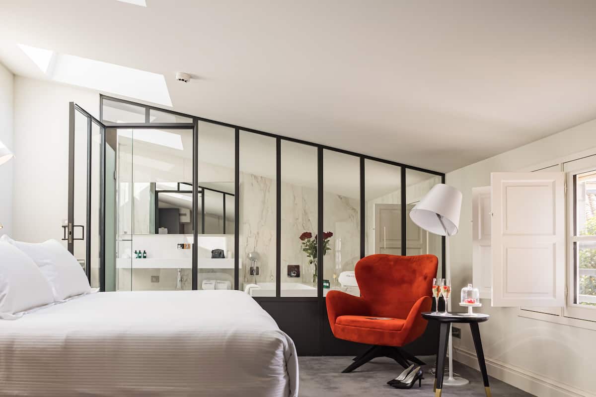 Quarto de hotel moderno com uma cama grande, uma poltrona vermelha, uma mesa pequena com copos e um espelho grande.