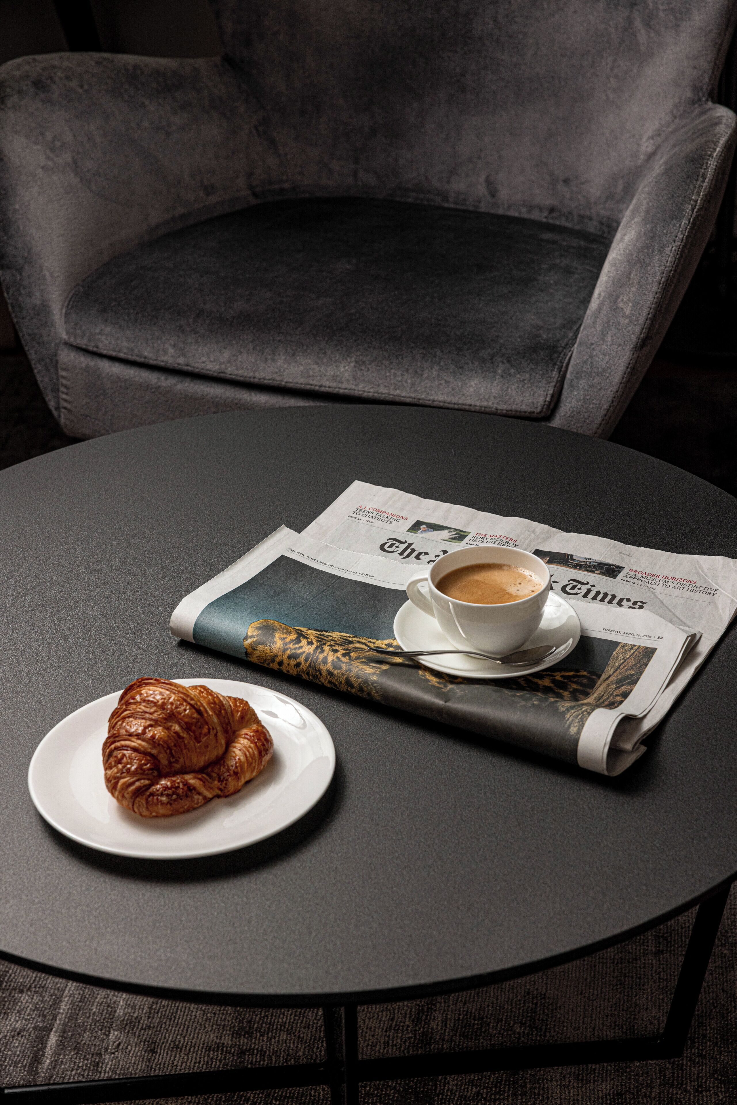 A round table with a newspaper, a cup of coffee, and a croissant.