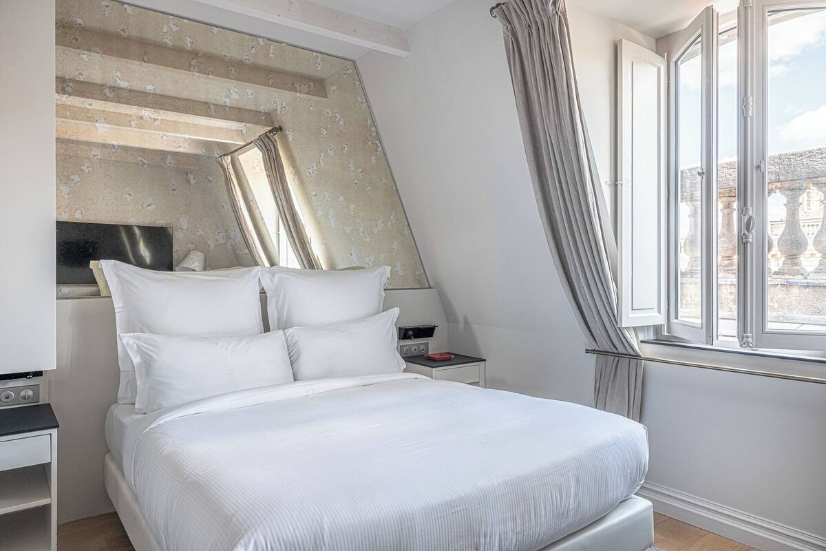 Quarto de hotel com cama, mesa de cabeceira, espelho, janela com cortinas e vista para o exterior.