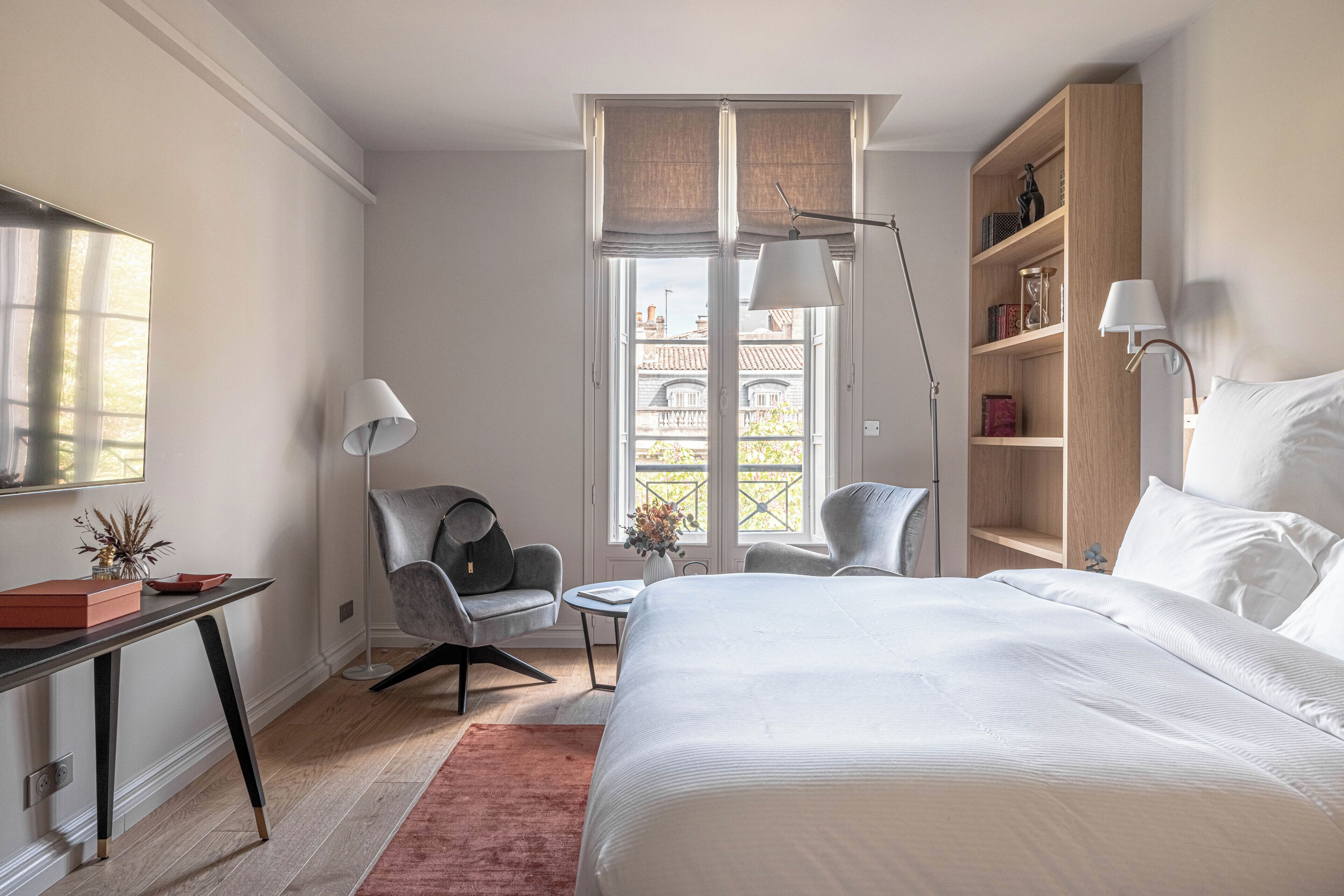 A hotel room with a bed, a desk, a chair, a bookshelf, and a window with a view.