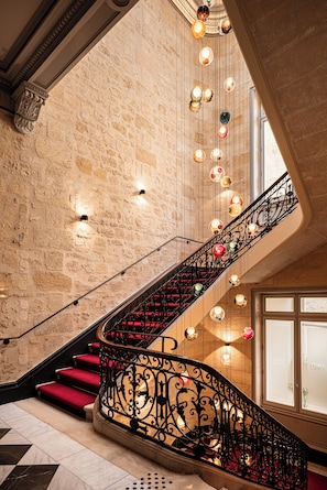 A grand staircase with ornate black railings, red carpeted steps, and decorative lighting fixtures against a stone wall backdrop. - Villas Foch (Bordeaux)