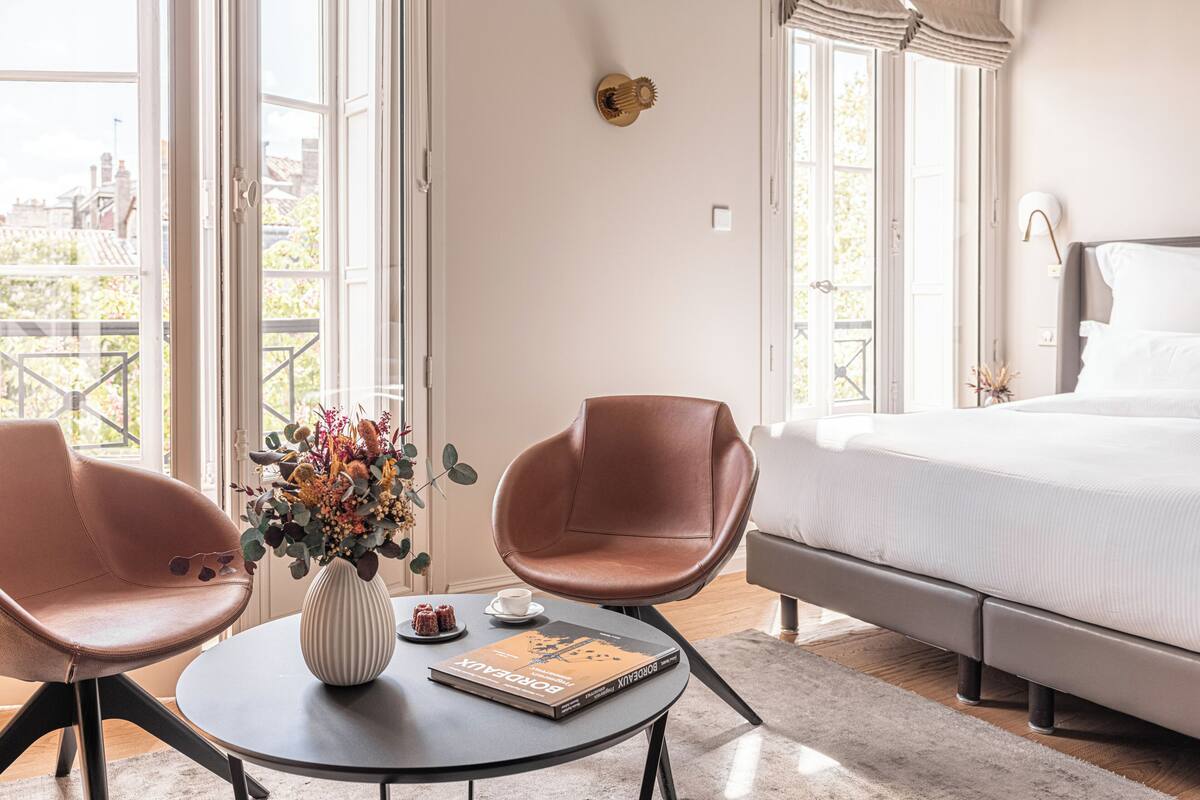 Quarto de hotel com cama, duas cadeiras, uma mesa com um vaso e um livro, e janelas amplas.
