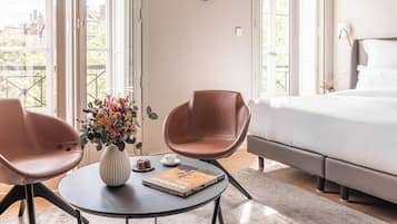 Ein Hotelzimmer mit einem Bett, zwei Stühlen, einem Tisch mit einer Vase und einem Buch sowie großen Fenstern.