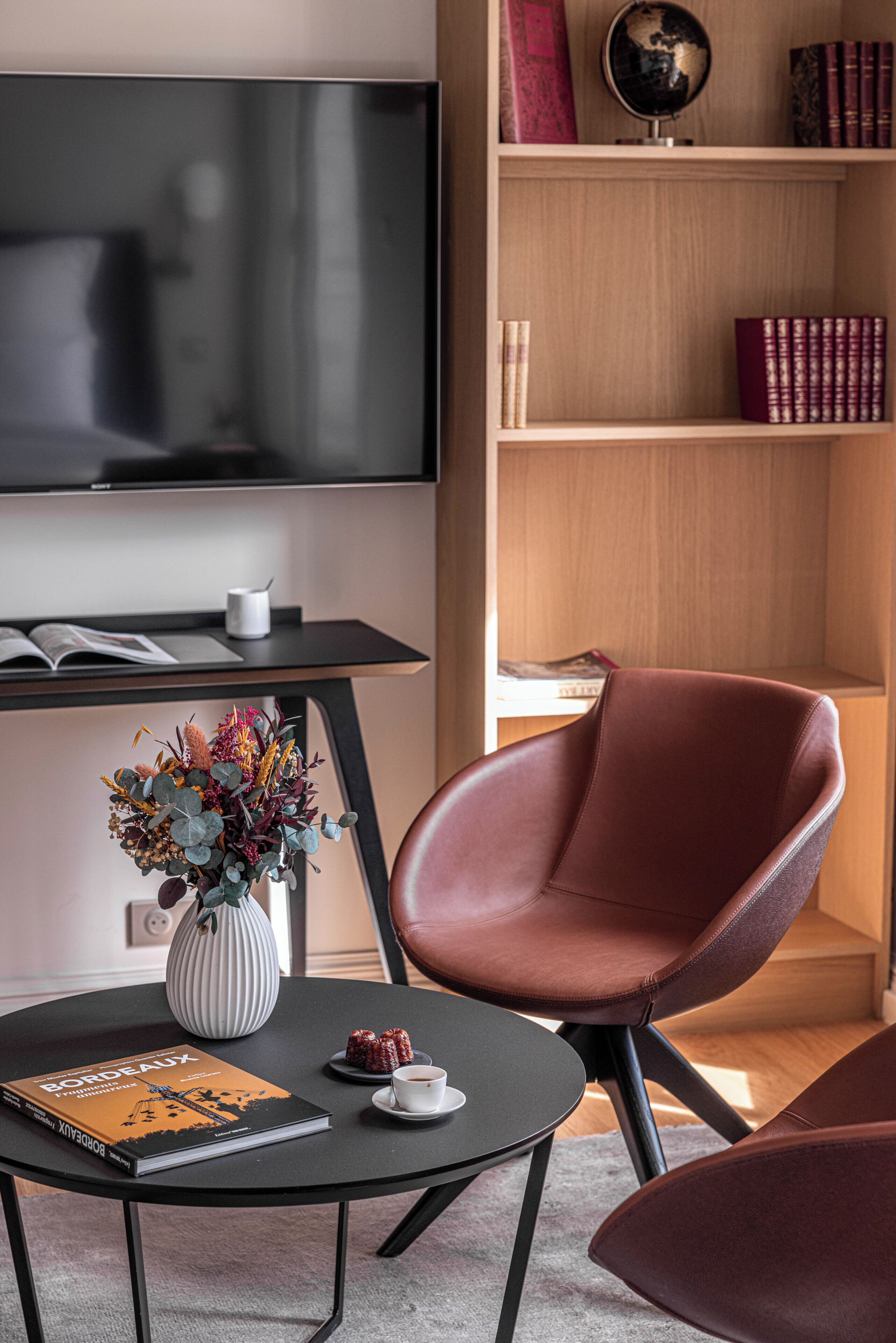 Uma sala de estar moderna com televisão de tela plana, estante de livros, poltrona de couro, mesa redonda com um livro e um vaso de flores.