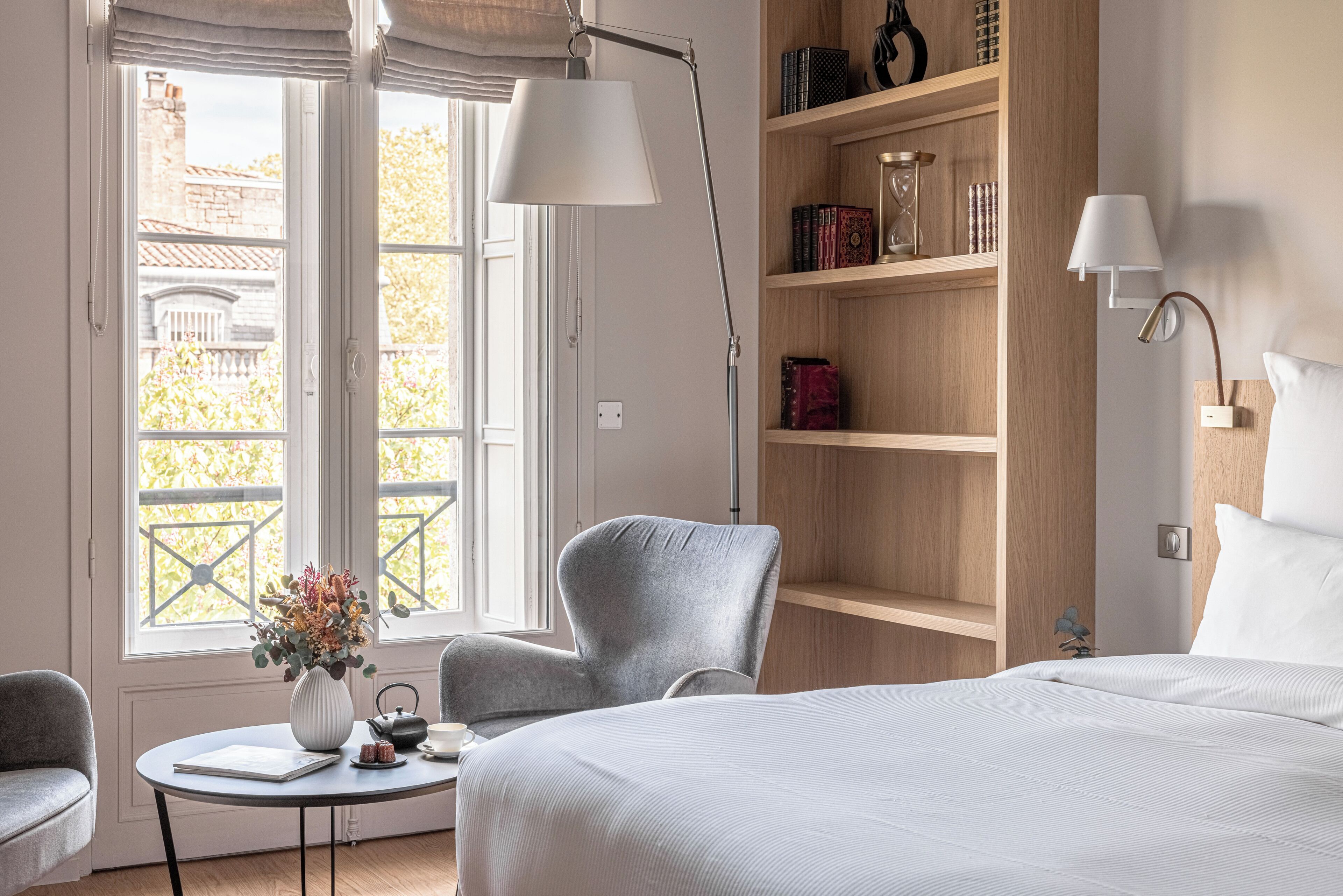 Habitación de hotel con cama, escritorio con lámpara, silla, estantería y vistas al exterior.