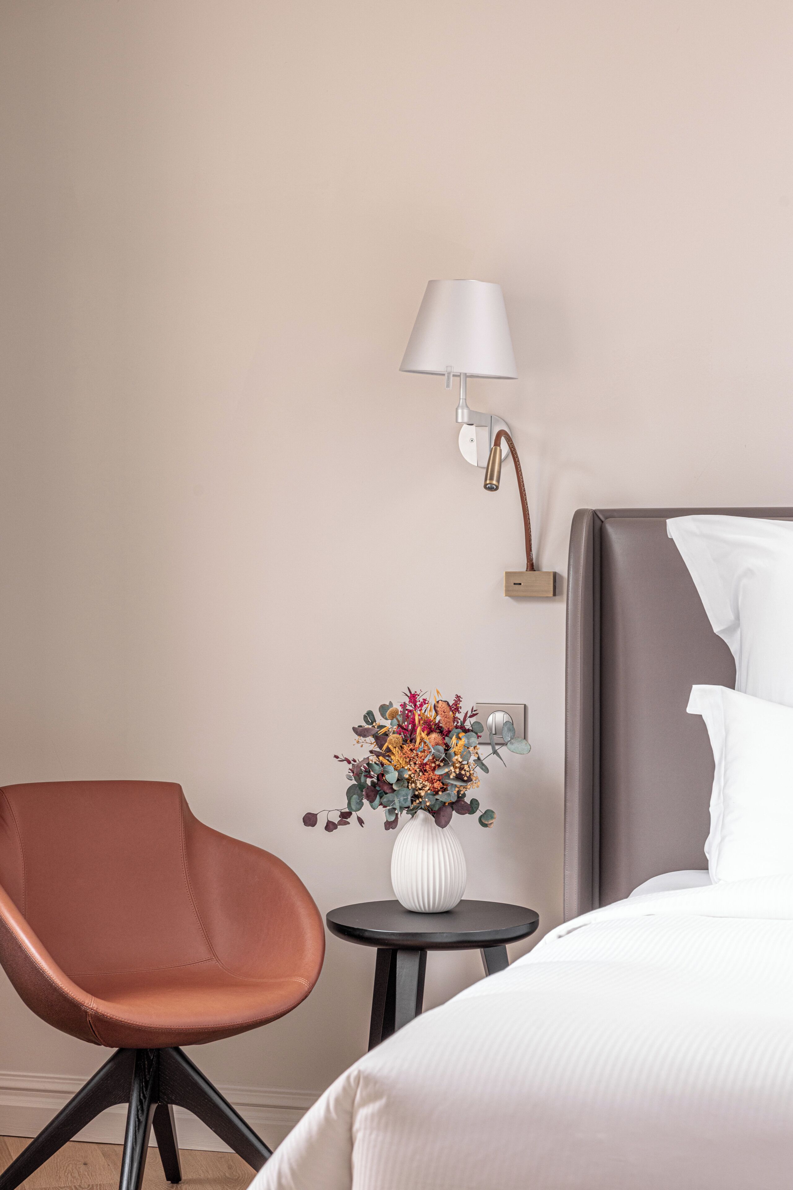 Una camera d'albergo con un letto, una sedia, un tavolino con un vaso di fiori e una lampada a parete.
