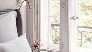 A bedroom with a bed, a bedside table with a book, a lamp, and a window with a view of buildings and trees. - Villas Foch (Bordeaux)