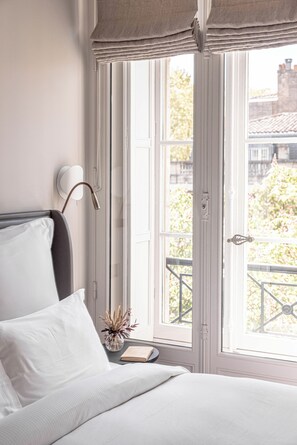 A bedroom with a bed, a bedside table with a book, a lamp, and a window with a view of buildings and trees. - Villas Foch (Bordeaux)