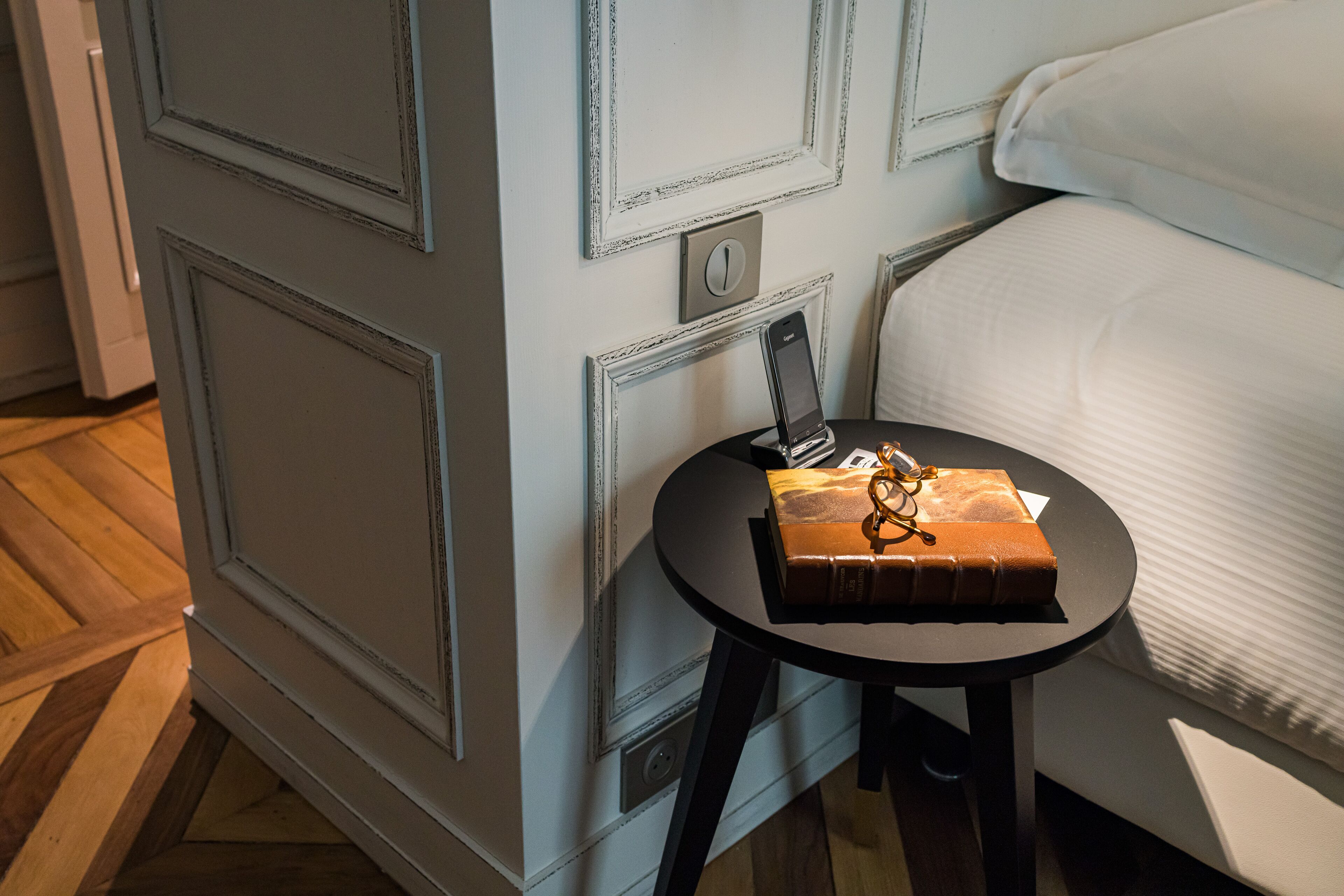 A room with a bed, a small table with a book and glasses, and a wall with a light switch and a thermostat.