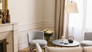 A room with a fireplace, two armchairs, a round table with a teapot and a vase of flowers, and a large window with curtains. - Villas Foch (Bordeaux)