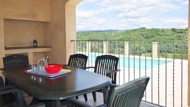 A dining area with a table and chairs, a red bowl, and a view of a swimming pool and greenery. - Quiet Holiday Home in Les Vans With Pool (Les Vans)