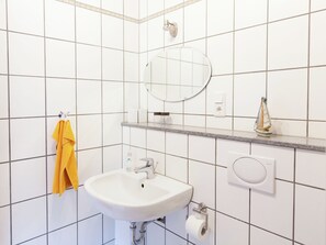 Kylpyhuone, jonka seinät on päällystetty valkoisilla laatoilla, ja jossa on valkoinen pesuallas, peili, keltainen pyyhe, pyyhekoukku, saippua-annostelija ja pieni koristeellinen purjevene.