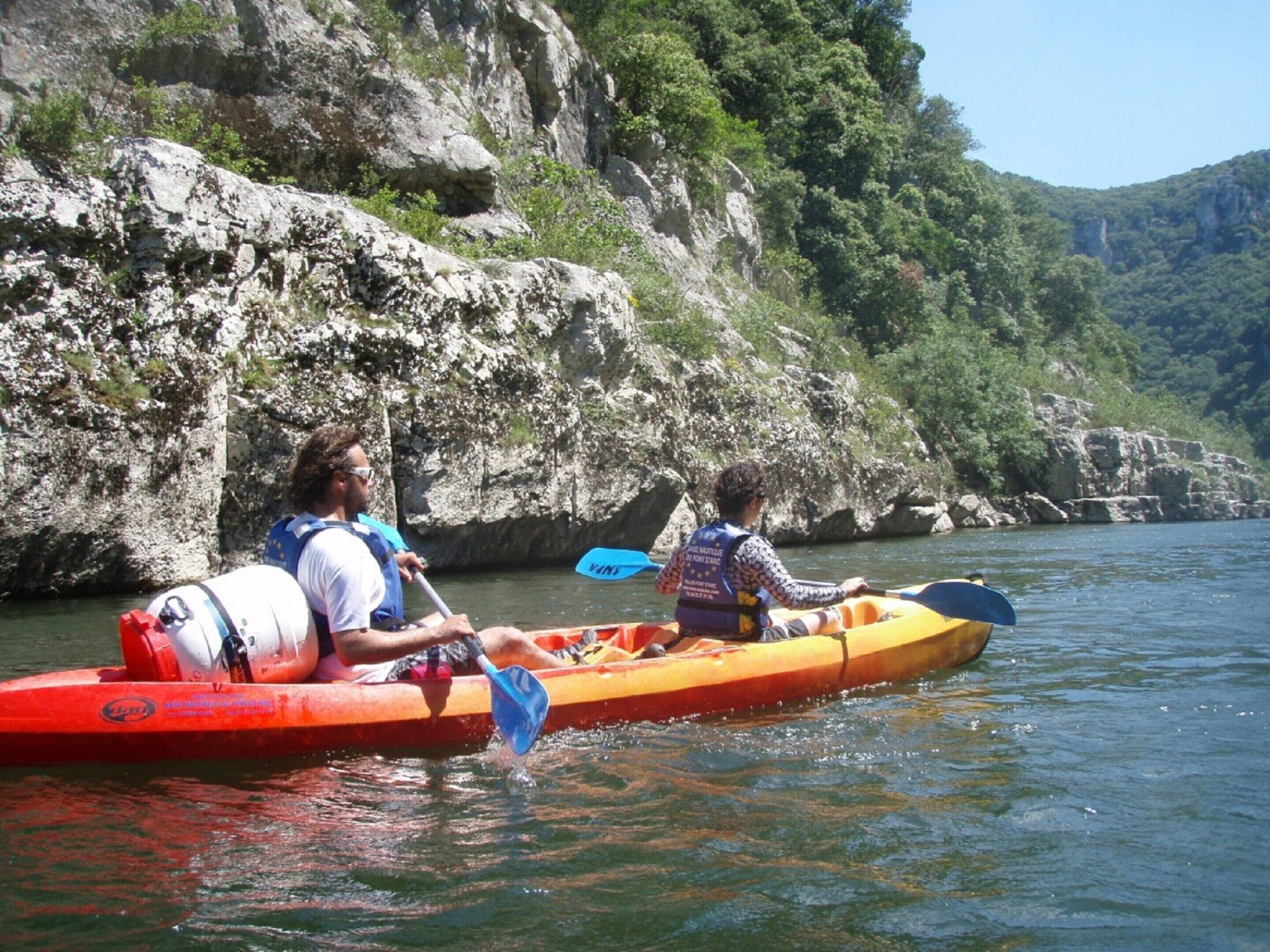 Two people kayaking on a river with rocky cliffs in the background.