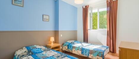 A bedroom with two beds, a window with curtains, and a lamp on a bedside table.