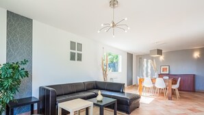 A modern living room with a black leather sofa, a small white table, and a dining area with a wooden table and white chairs. - Charming Villa Near Les Vans (Les Vans)