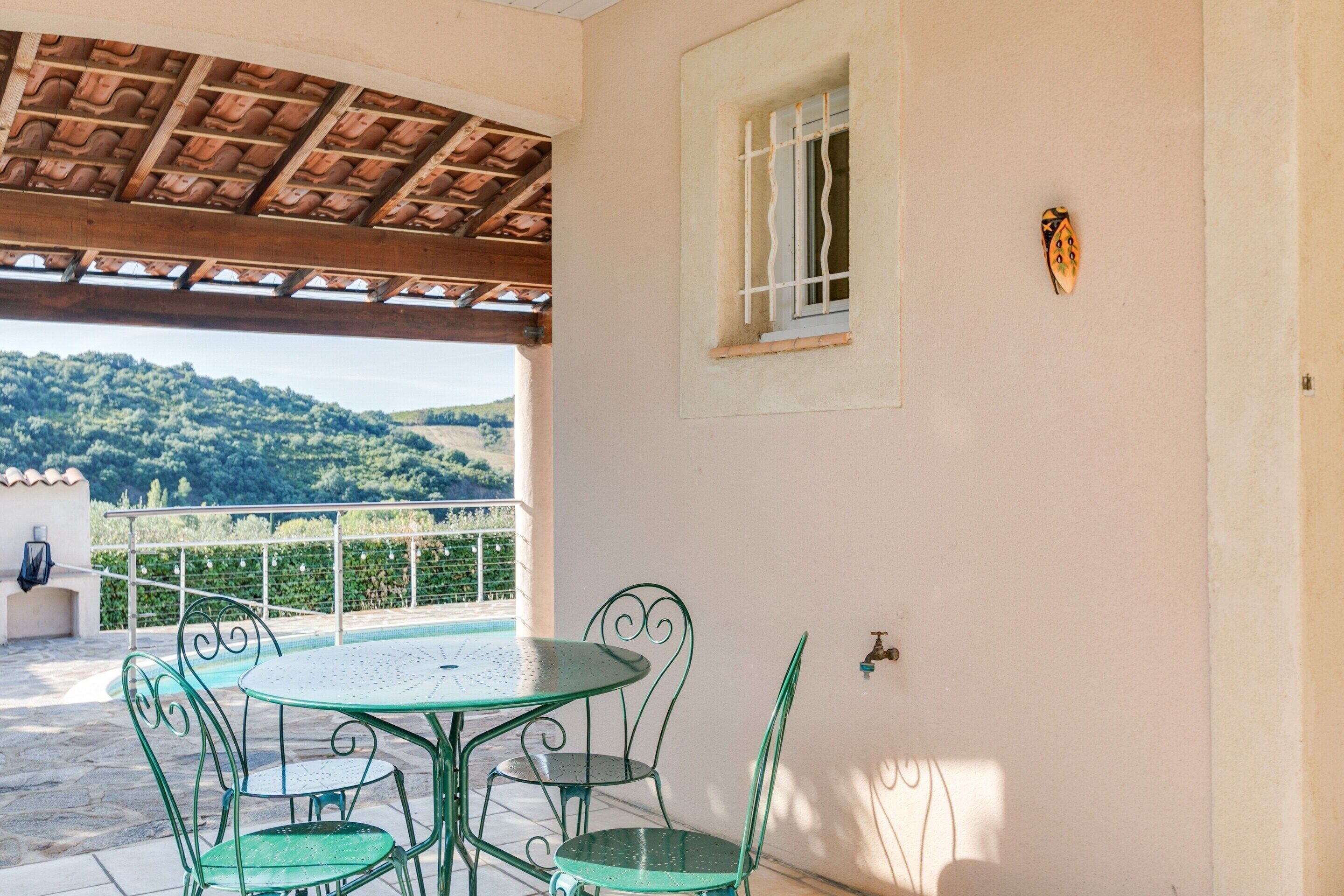 A patio with a round table and four chairs, a window with a decorative wall piece, and a view of a hilly landscape.