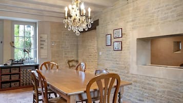 A dining area with a wooden table and chairs, a chandelier, stone walls, and a window with a view of greenery.