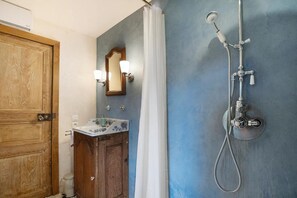 A bathroom with a wooden door, a sink with a floral design, a mirror with wall sconces, and a shower with a handheld showerhead. - Manor in Normandy With Spa and Pool (Ponts sur Seulles)