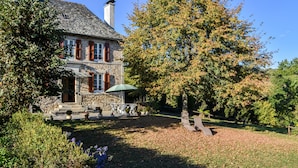 Uma casa de pedra com chaminé, uma árvore com folhas de outono e um jardim com um banco.